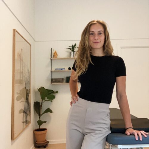 Camilla standing by the treatment couch in the osteopathy clinic room. DA: Camilla stående ved behandlingsbriksen i et behandlerrum på en osteopati klinik.